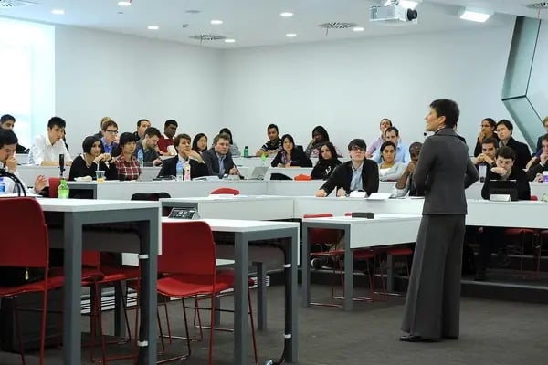 Professorin hält eine Vorlesung in einem modernen Hörsaal mit weißen Wänden, roten Stühlen und Studierenden, die aufmerksam zuhören.