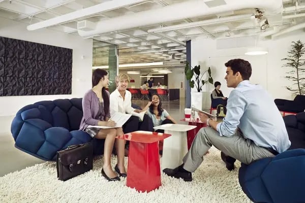 Studierende sitzen in einer modernen Lounge mit stilvollen blauen Sofas und roten Tischen und führen eine Diskussion in entspannter Atmosphäre.