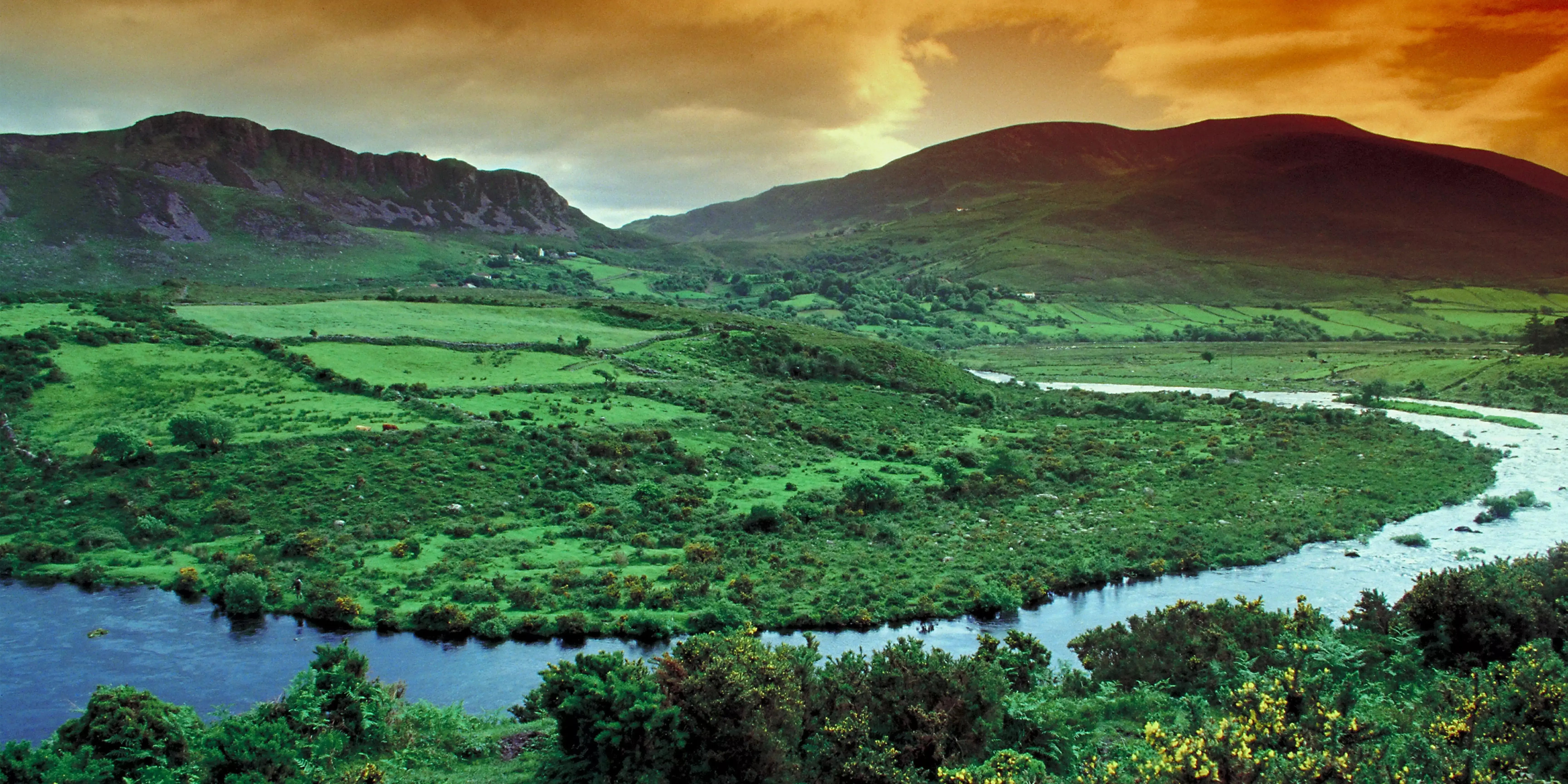 Grüne Hügellandschaft mit Flusslauf bei Sonnenuntergang. Malerische Naturkulisse, typisch für die Landschaft Irlands.