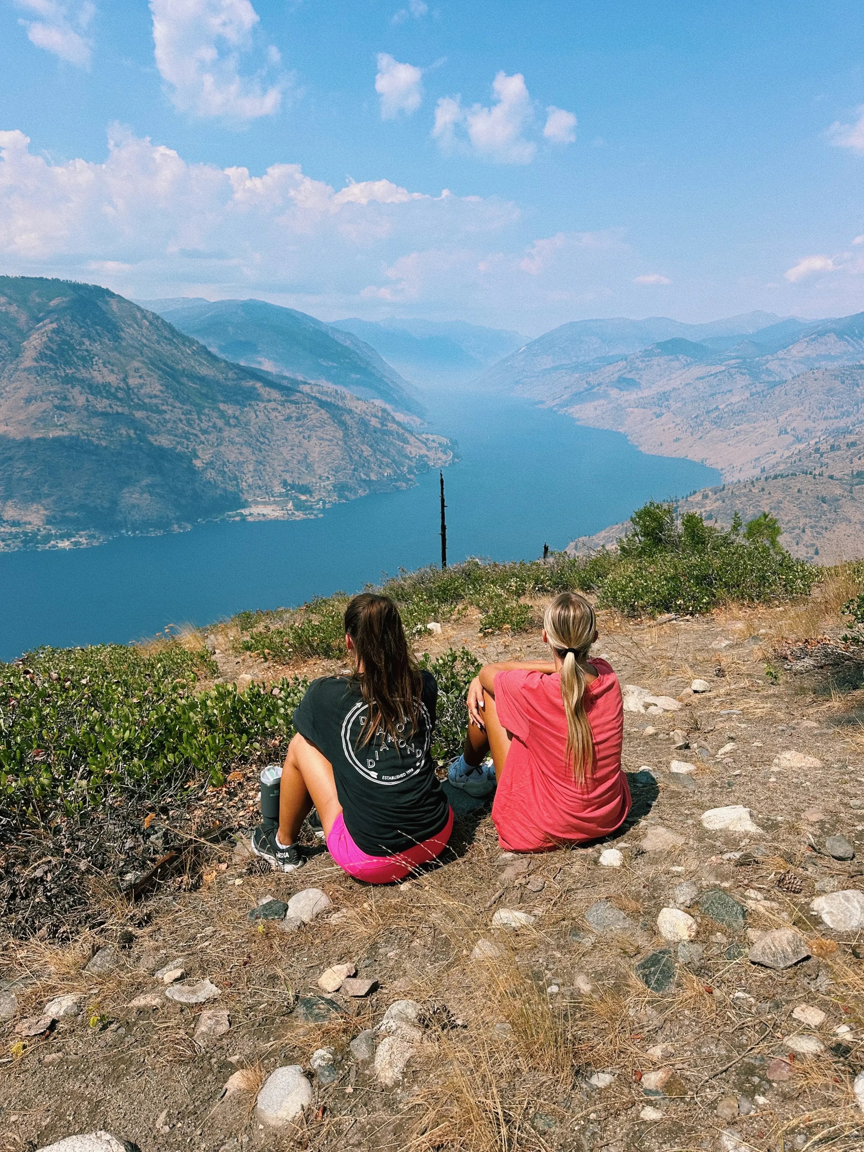 Zwei Mädchen sitzen an einem Hang in der Sonne und blicken über einen Canyon mit Fluss